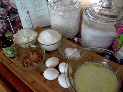 overhead view of glass bowls and jars filled with ingredients for making chocolate mississippi mud cake such as eggs, sugar, flour, butter and cocoa powder all on a brown wooden table.