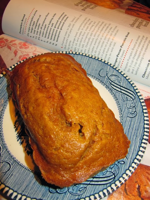 overhead view of a whole pumpkin bread loaf on a white platter with a light blue decorative rim set on a cook book open to a pumpkin bread