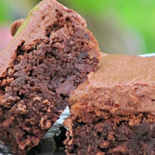 Two fudgy chocolate brownies from Joy the Baker with malt-flavored chocolate frosting sit with one at an angle resting on the other on white parchment paper against a green background
