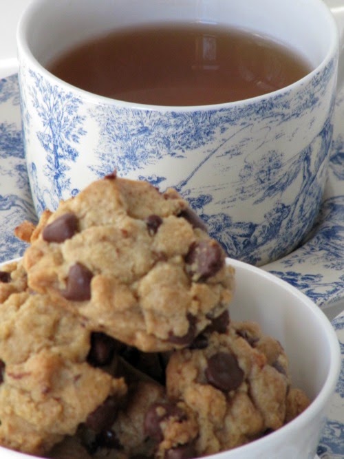Several Browned Butter Chocolate Chip Cookies in a white bowl in front of a blue and white cup full of coffee and matching saucer.