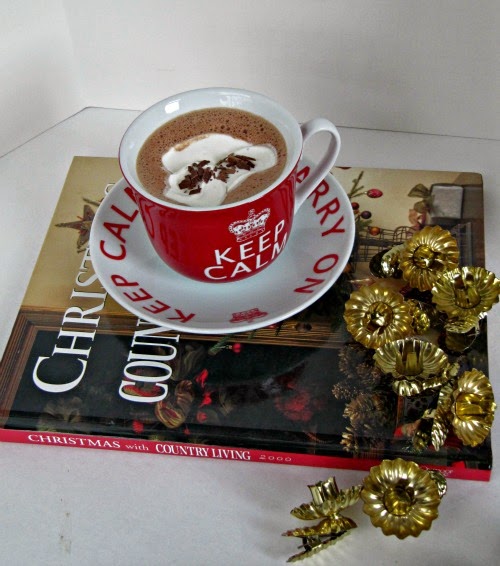 overhead view of a cup of gingerbvread cocoa in a red china cup on a red and white saucer that says "Keep Calm" sitting on a red christmas book on a white table with gold decorations scattered to the right of the cup and saucer spilling over the book onto the table.