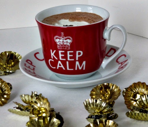 gingerbread hot cocoa in a large red mug that says "Keep Calm" on a white saucer on a white table surrounded with gold christmas decorations