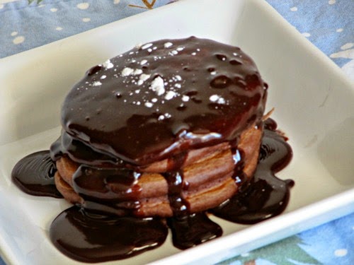 Stack of chocolate cocoa pancakes dripping with fudge sauce and a sprinkle of powdered sugar on a white plate.