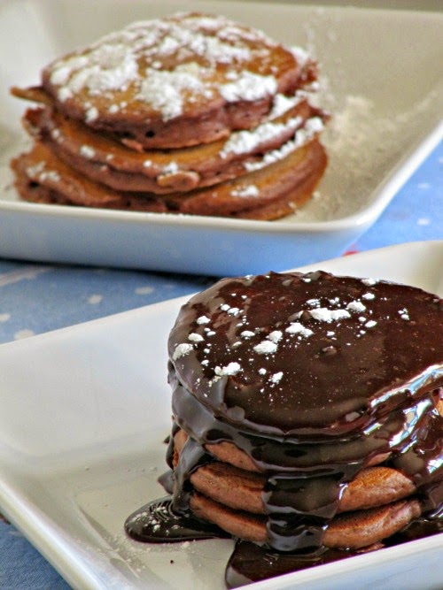 Two stacks of rich chocolate pancakes with chocolate fudge sauce in foreground and with powdered sugar in background