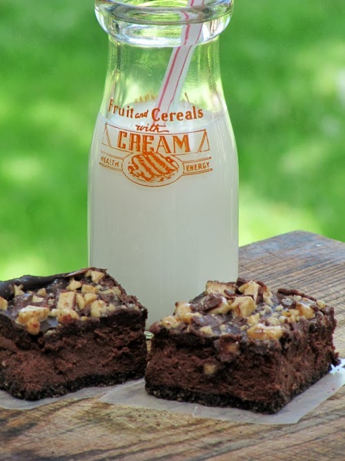 Two dark chocolate cheesecake squares with chopped toffee and chocolate glaze on top sitting on a wood board in front of a white old-fashioned glass milk bottle with red lettering. The cheesecake and milk bottle are against a mottled green background.