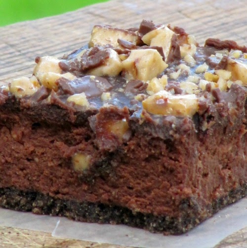 Close-up photo of a dark chocolate cheesecake square with chopped toffee and chocolate glaze on top sitting on a white plate against a mottled green background.