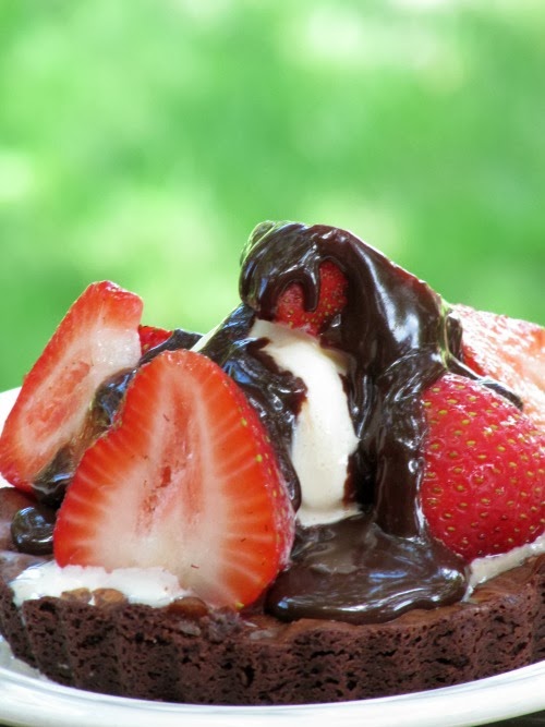 side view of a round brownie tart with sliced strawberries on top surrounding a scoop of vanilla ice cream and covered with hot fudge sauce. The tart is against a green background