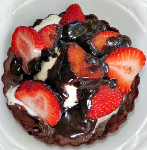 close up top view of round fresh fruit  brownie tart sundae covered with vanilla ice cream, chocolate fudge sauce and sliced strawberries on a white plate.