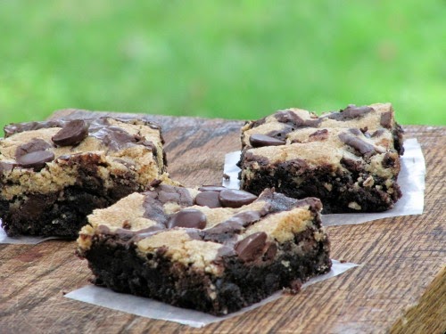Chocolate Chip Cookie Brownies sitting on a brown table on pieces of parchment paper, each with a chocolate brownie base and chocolate chip cookie top layer.