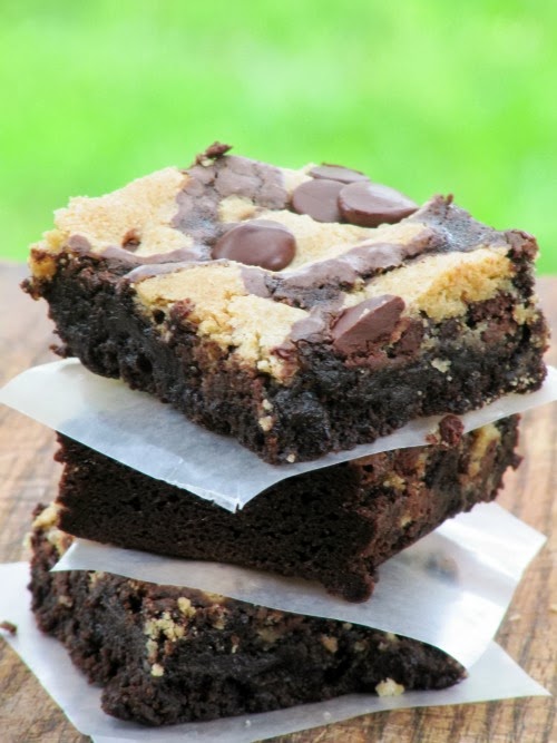 Stack of three fudgy chocolate chip cookie brownies with a chocolate chip cookie layer full of melted chocolate chips. The stack is sitting on a brown table. 