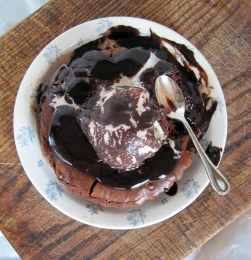 overhead view of a round fudge brownie tart from Southern Living Magazine covered with vanilla ice cream and hot fudge sauce on a white plate with blue flower decorations. A silver spoon rests on top of the tart and the platter is set on a wood serving board on top of crumpled white parchment paper.