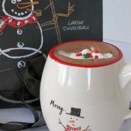 closeup of a eed ringed white ceramic mug filled with hot cocoa topped with whipped cream and red and green holiday candies on a white table along with a black chalkboard with a white chalk snowman drawing.