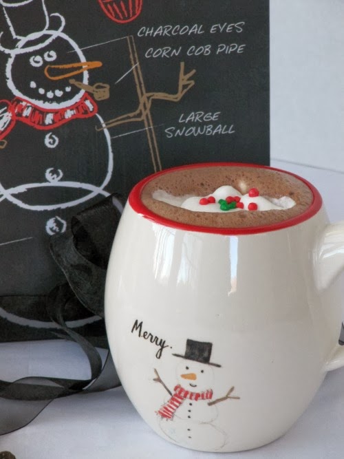 closeup of a eed ringed white ceramic mug filled with hot cocoa topped with whipped cream and red and green holiday candies on a white table along with a black chalkboard with a white chalk snowman drawing.