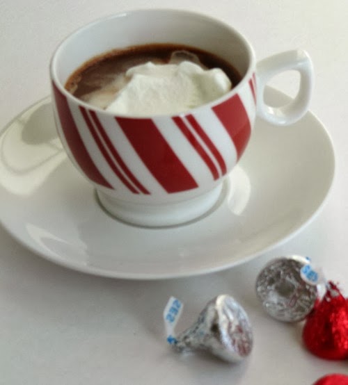 A red and white striped cup filled with creamy hot cocoa topped with whipped cream sits on a white saucer of a white background that is scattered with hershey's silver and red kisses candies