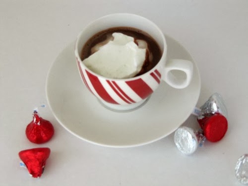 An overhead view of a peppermint red and white striped cup of Creamy Hot Cocoa sits on a white saucer on a white surface that has hershey's silver and red chocolate kisses scattered on it.