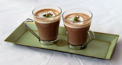 distance angled view of two glass mugs of Bailey's Hot Chocolate made with dark chocolate, Bailey's Irish cream and topped with whipped cream and chopped chocolate. The mugs are on a green tray against a light gray background.