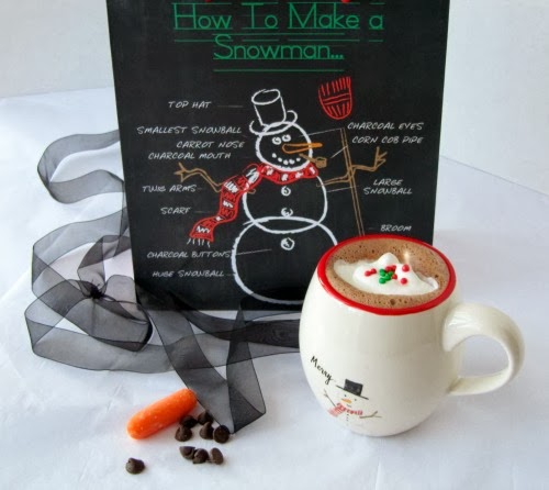 Red ringed white ceraic mug filled with Green & Black's Luxury Cocoa topped with whipped cream and red and green holiday candies on a white table along with a black chalkboard with a snowman drawing along with holiday decorations.