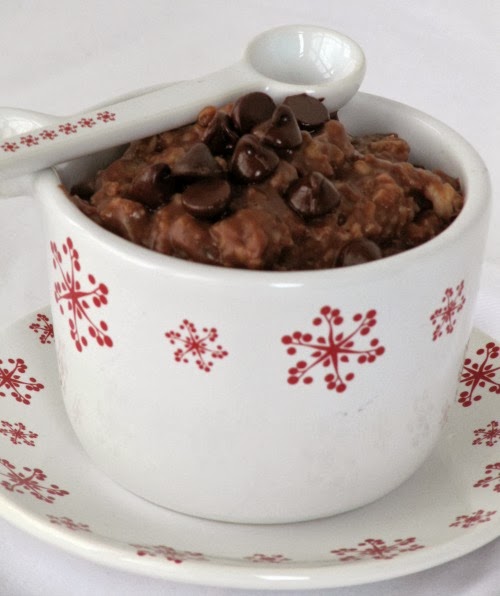 white ceramic cup with red snowflake decorations filled with chocolate chocolate chip oatmeal on a saucer with the same decorations. A matching white spoon is resting along the top of the mug.