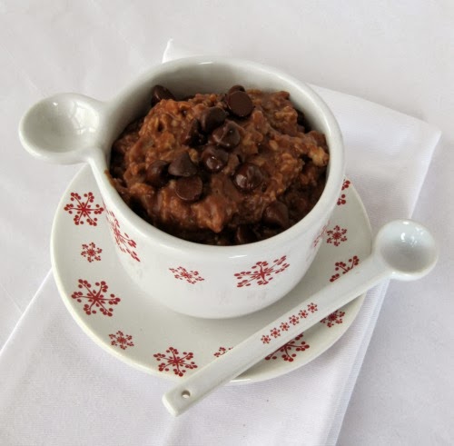 top view of a white ceramic cup with red snowflake decorations filled with chocolate chocolate chip oatmeal on a saucer with the same decorations. A white measuring spoon is resting along the top of the mug. Mug and saucer are on a white cloth napkin on a white table cloth.