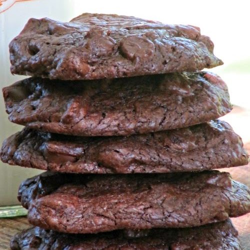 Close up of a stack of five large chocolate cookies with chocolate chips inside sitting on a brown table with a glass milk bottle peeking out from the left side of the photo.