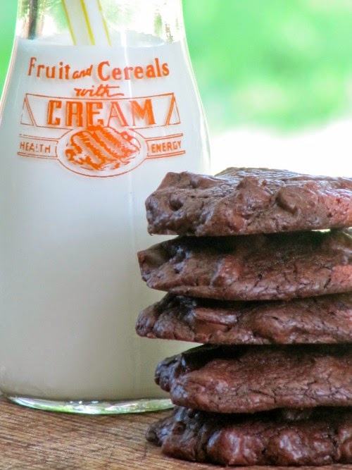 Stack of five large outrageous chocolate chocolate cookies studded with chocolate chips on a brown table with a glass milk bottle with red writing on it is behind them, in front of a green background.