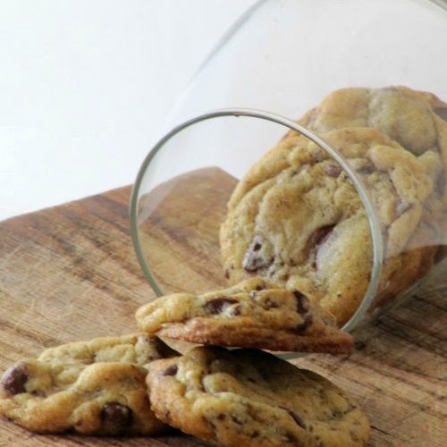 a pile of the best chocolate chip cookies ever tumbling out of a glass jar onto a wood tabletop