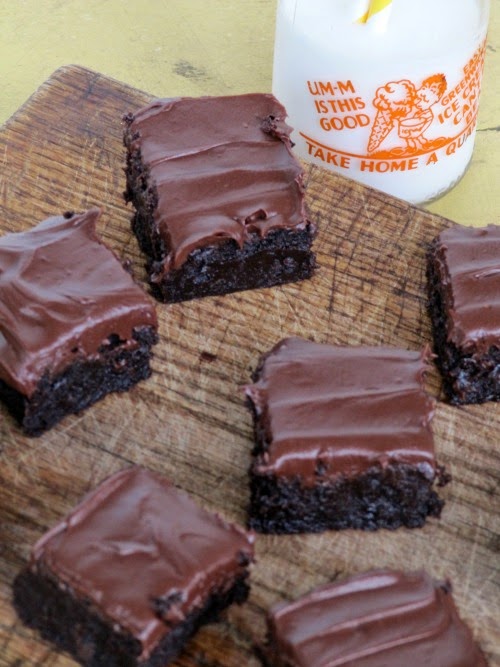 several fudgy double chocolate bourbon brownies with hershey's chocolate frosting are sitting on a wooden cutting board in front of a glass bottle of milk.