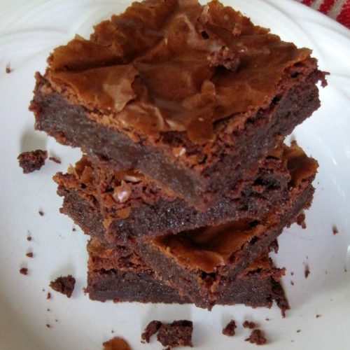 A stack of three chocolate Connie's PMS brownies with crinkly tops on a white plate surrounded with crumbs