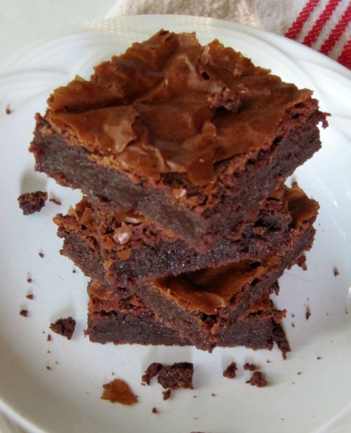 A stack of three chocolate Connie's PMS brownies with crinkly tops on a white plate surrounded with crumbs