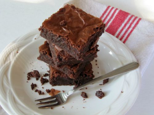 a white plate is holding a stack of three dark chocolatey fudge Connie's PMS brownies with crispy tops surrounded by brownie crumbs and a fork is in front of the stack.