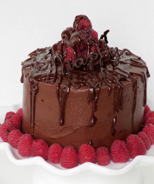 Round chocolate frosted chocolate buttermilk cake with whipped coca frosting and has chocolate drizzle all over and a centerpiece of fresh raspberries in a mound with the word happy written in chocolate in front of the raspberreis and a ring of fresh raspberries around the bottom of the cake.