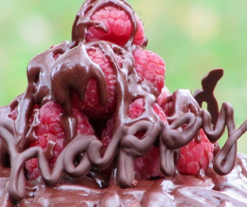 closeup of a chocolate frosted chocolate buttermilk cake-top with a pile of fresh raspberries covered in a drizzle of chocolate glaze with the word "happy" written in chocolate.