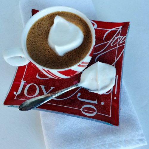 Top view of a roundpeppermint striped mug filled with hot chocolate and a dollop of whipped cream on a red square tray with 'Joy" written in white letters on it. Tray has a silver serving spoon filed with whipped cream to the left of the mug. Tray is on a white tablecloth