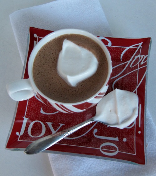 Top view of a round peppermint striped mug filled with hot chocolate and a dollop of whipped cream on a red square tray with 'Joy" written in white letters on it. Tray has a silver serving spoon filed with whipped cream to the left of the mug. Tray is on a white tablecloth.