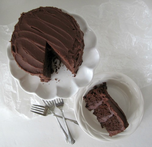 Top view of dark chocolate blackout  layer cake on a white cake stand with a slice of chocolate cake ona plate in front of the layer cake.