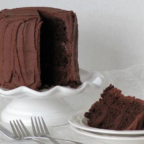 Three-layer "Mom's" blackout chocolate cake with chocolate frosting on a white cake stand with one slice of the cake on a stack of white plates