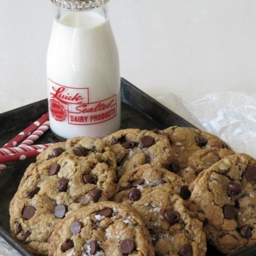 Brown tray of chocolate chip cookie with a glass milk bottle with red writing on it filled with milk on the tray next to the cookies and red and white straws on the tray near the milk bottle.
