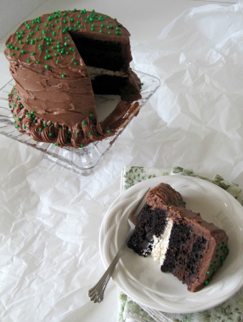 a piece of chocolate layer cake made with guiness stout beer with cream cheese and chocolate frosting between the layers is on a white plate. Behind the plate is a chocolate frosted round layer cake
