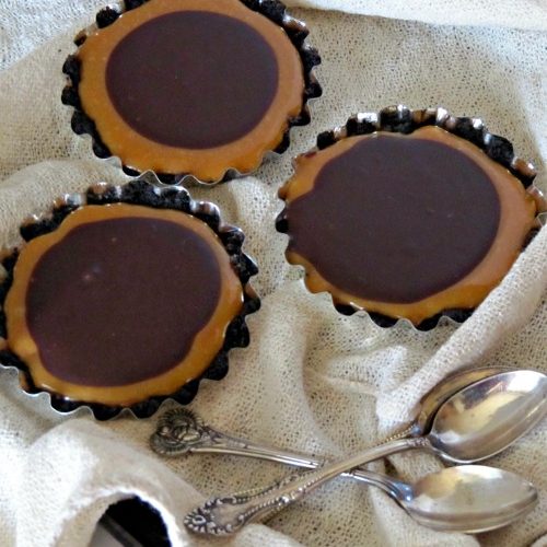 top view of three little chocolate caramel turtle tarts on a white table cloth with 2 silver serving spoons criss-crossed in front of them