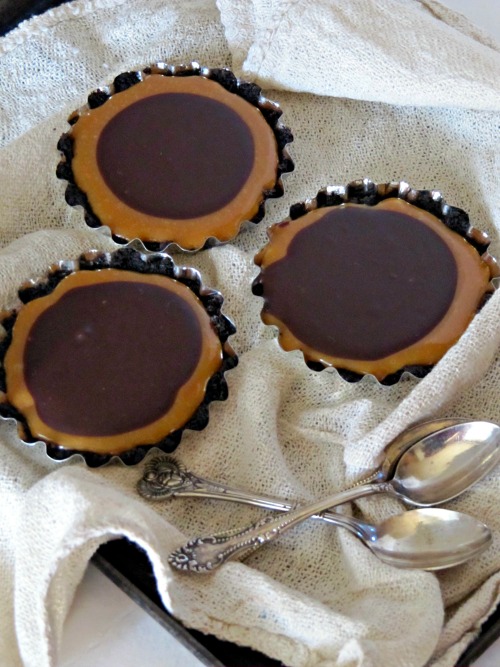 three chocolate caramel turtle tarts in metal tart tins on a crumpled white table cloth with two silver spoons criss-crossed in front of them
