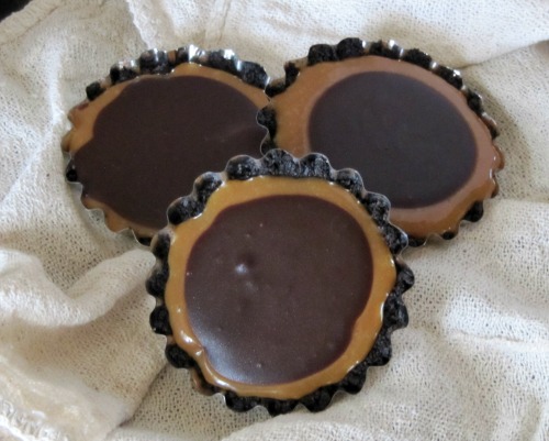 top view of three little chocolate caramel turtle tarts on a white table cloth with 2 silver serving spoons criss-crossed in front of them