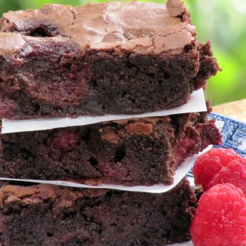 closeup of a stack of three fudgy raspberry brownies separated by white parchment paper in between on a blue and white plate with fresh raspberries at their side.