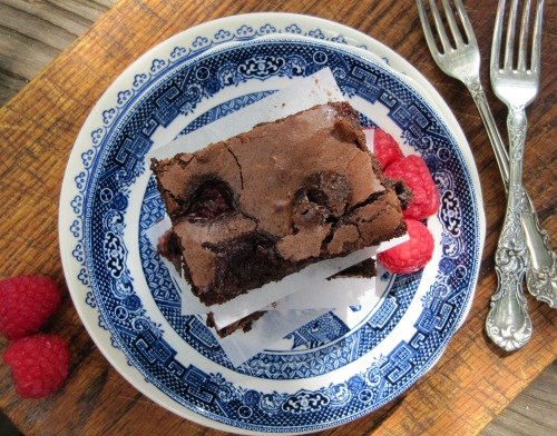 Overhead view of a large dark chocolate brownie filled with raspberries on a blue and white plate with a few fresh raspberries on the side