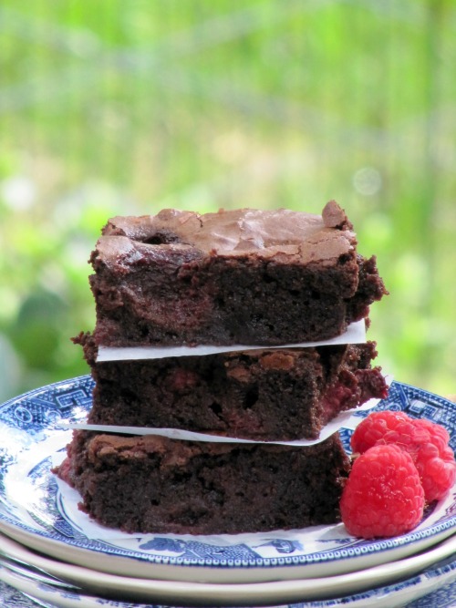 A blue plate has a stack of rich chocolate fudgy raspberry brownies with white parchment paper separating them. Three fresh raspberries are also on the plate to the side of the brownie.s