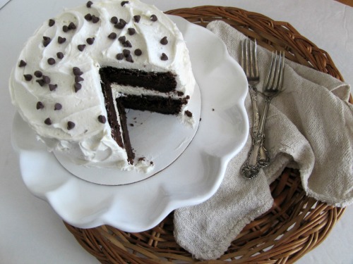Two-layer dark chocolate devils food cake with white buttercream icing and chocolate chip garnish on a white cake stand with a slice of the cake on a white plate in front.