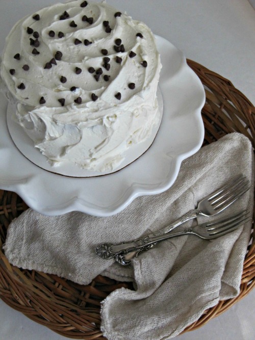 Whole round devils food dark chocolate cake with white buttercream frosting decorated with chocolate chips on a white cake stand.