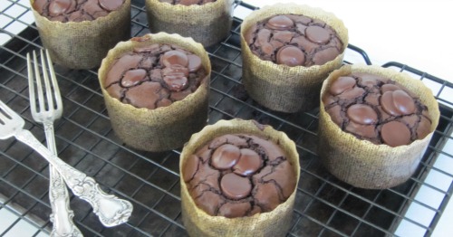 Top view of chocolate brownie muffins studded with chocolate chips wrapped in parchment paper sitting on a dark brown tray on a cooling rack with two silver forks criss-crossed on the left side of the tray