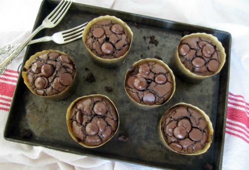 Top view of chocolate brownie muffins studded with chocolate chips wrapped in parchment paper sitting on a dark brown tray on a cooling rack with two silver forks criss-crossed on the left side of the tray. Tray is on a white table cloth with a red stripe down the middle