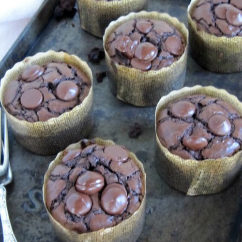 Chocolate brownie muffins studded with chocolate chips wrapped in parchment paper sitting on a dark brown tray on a white table