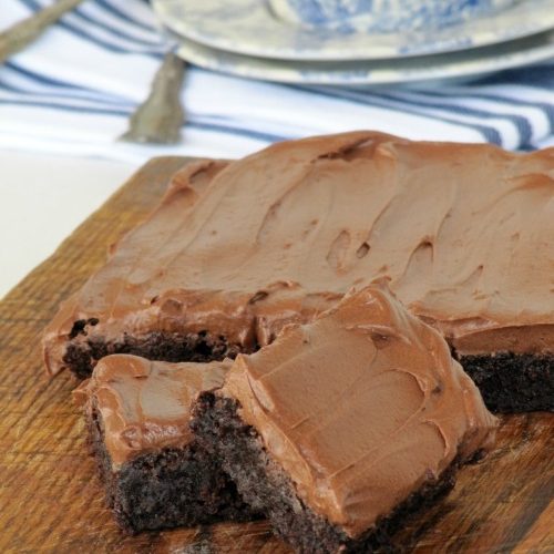 Slab of frosted fudgy cocoa brownies with two cut brownie squares in front on a wooden cutting board